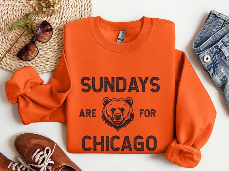 K&ouml;nnte beinhalten: Orangefarbener Pullover mit einem schwarzen grafischen Aufdruck, der "Sundays are for Chicago" mit einer B&auml;renillustration sagt.