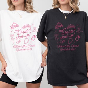 May include: White and black t-shirts with a pink graphic design. The design includes the text "me? a bride? shut up!" along with illustrations of a crown, ring, and other bridal-themed elements. The text "Future Mrs. Smith Scottsdale 2025" is also included.