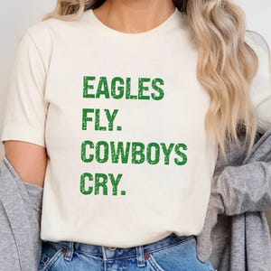 May include: Cream-colored t-shirt with the text "EAGLES FLY. COWBOYS CRY." in green, distressed lettering. The shirt is a casual, short-sleeved style, suitable for everyday wear. The overall theme is sports and team spirit.