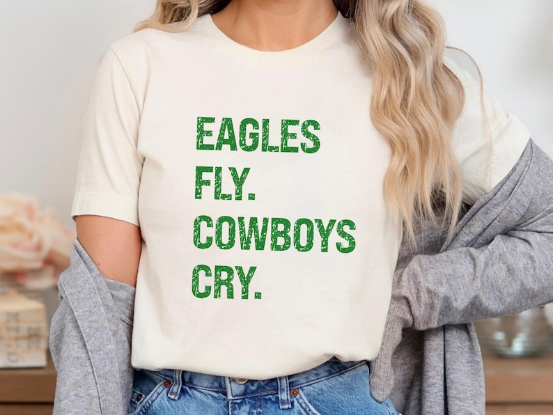 May include: Cream-colored t-shirt with the text "EAGLES FLY. COWBOYS CRY." in green, distressed lettering. The shirt is a casual, short-sleeved style, suitable for everyday wear. The overall theme is sports and team spirit.