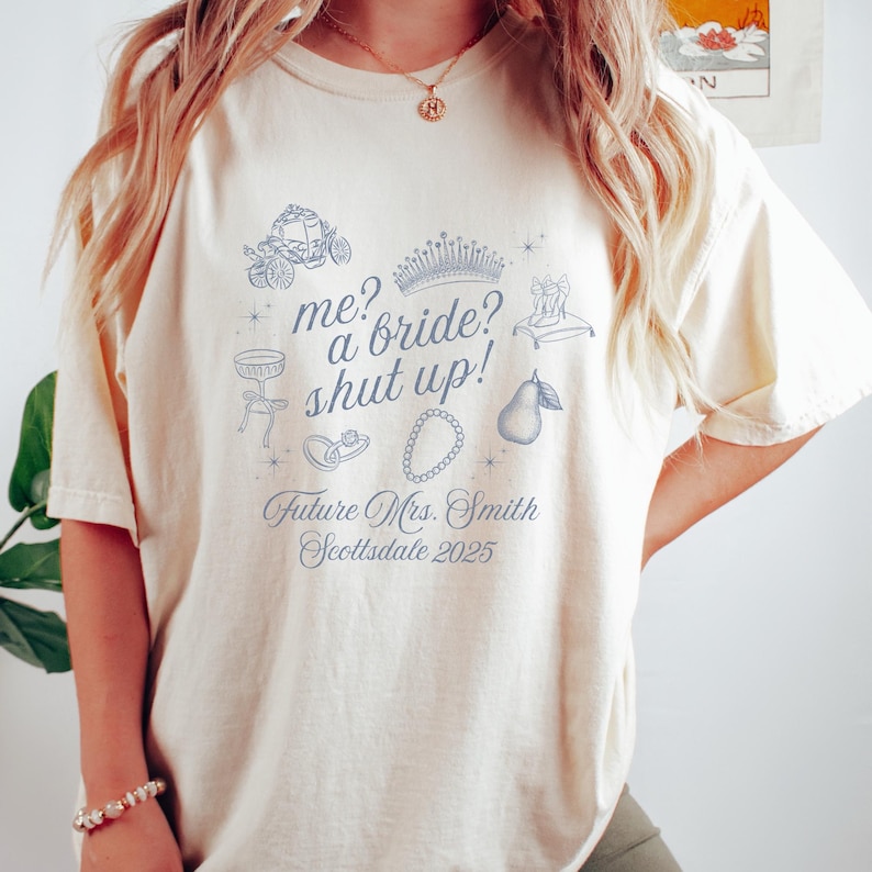 May include: Cream-colored t-shirt with a blue graphic design. The design includes a carriage, crown, and the text "me? a bride? shut up!" along with "Future Mrs. Smith Scottsdale 2025".