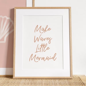 May include: A framed print with the text "Make Waves Little Mermaid" in a light brown cursive font on a white background. The frame is a light wood colour.