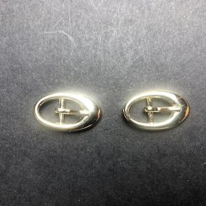 Peut inclure: Deux boucles de ceinture ovales dorées. Chaque boucle a une barre centrale et un cadre arrondi. Les boucles sont posées sur un fond sombre, soulignant leur éclat métallique. Idéales pour les ceintures ou accessoires.