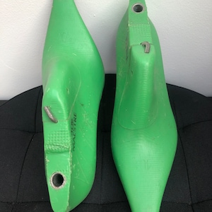 May include: Two green shoe lasts, one marked "10 1/2" and the other "46". The lasts are made of a solid material and have a pointed toe shape. They have metal hardware and are designed for shoemaking.