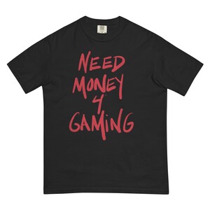 May include: Black t-shirt featuring the red text "NEED MONEY 4 GAMING". The text is in a handwritten style. The t-shirt has a crew neck and short sleeves. The material looks soft and comfortable.