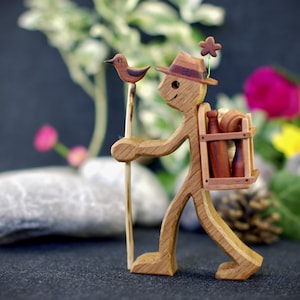 Könnte beinhalten: Eine Holzfigur eines Menschen, der eine kleine Holzkiste auf dem Rücken trägt. Die Person trägt einen Hut und hat einen Vogel auf der Schulter sitzen. Die Figur steht auf einem Holzsockel.