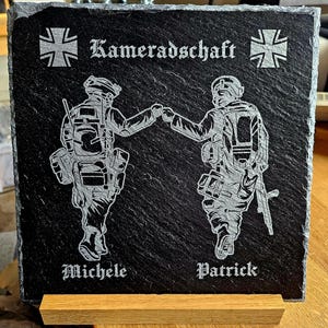 Puede incluir: Una placa de pizarra negra con dos figuras de soldados en una pose militar, con el texto "Kameradschaft" y los nombres "Michele" y "Patrick" grabados en la placa.