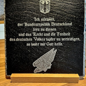 Op de afbeelding: Een zwarte leisteenplaat met een witte adelaar en tekst die luidt "Ich schwöre, der Bundesrepublik Deutschland treu zu dienen und das Recht und die Freiheit des deutschen Volkes tapfer zu verteidigen, so wahr mir Gott helfe."