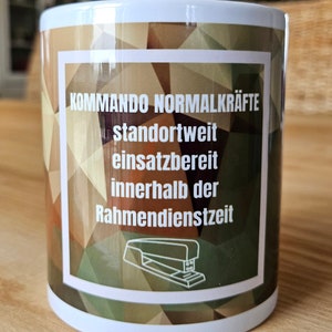 Könnte beinhalten: Weißer Keramikbecher mit einem geometrischen Tarnmuster in Braun-, Grün- und Beigetönen. Der Becher zeigt deutschen Text und eine Strichzeichnung eines Tacker in einem weißen rechteckigen Rahmen.