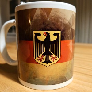Könnte beinhalten: Weiße Keramiktasse mit einem Vintage-Design der deutschen Flagge. Die Flagge zeigt die Farben Schwarz, Rot und Gold mit einem stilisierten schwarzen Adler in der Mitte.