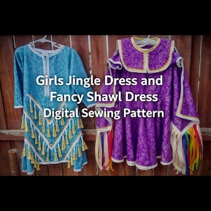 May include: Two dresses, a Girls Jingle Dress and a Fancy Shawl Dress, hanging on a wooden background. The blue Jingle Dress has gold fringe. The purple Fancy Shawl Dress has colorful ribbons.