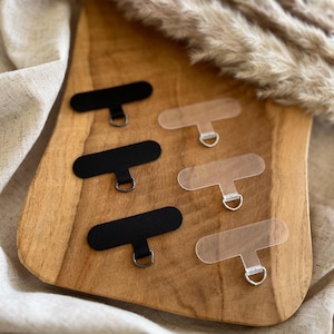 Könnte beinhalten: Sechs Klebehaken, vier schwarze und zwei transparente, mit Metallringen. Die Haken befinden sich auf einer Holzoberfläche.