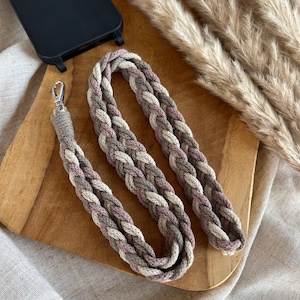Könnte beinhalten: Geflochtenes Handy-Lanyard in Grau-, Mauve- und Cremetönen mit silberfarbenem Verschluss. Das Lanyard liegt auf einem Holzschneidebrett, neben einer schwarzen Handyhülle und getrocknetem Ziergras.