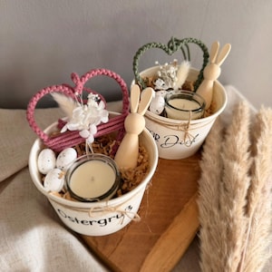 Könnte beinhalten: Zwei weiße Metalleimer mit Osterdekorationen. Jeder Eimer enthält eine Kerze, einen Holzhasen, dekorative Eier und Trockenblumen. Ein Eimer hat ein herzförmiges Makramee in Rosa, der andere in Grün. Die Eimer stehen auf einem Holztablett.