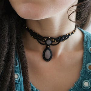 May include: Black macrame necklace with a teardrop-shaped pendant. The necklace features a woven design with small gold-colored beads. The pendant is a light gray oval stone. The necklace is worn against a teal-colored top.