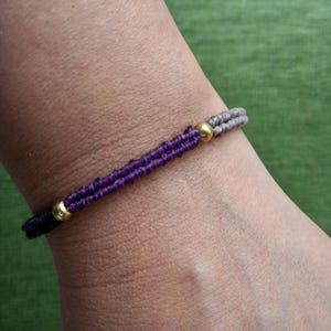 Puede incluir: Una pulsera de macramé con una sección central morada, flanqueada por secciones grises y negras. La pulsera está adornada con dos cuentas doradas. La pulsera se lleva en una muñeca.