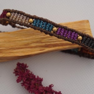 Puede incluir: Una pulsera de macramé con un diseño tejido que presenta secciones de color alternas. La pulsera incluye secciones de borgoña, beige, turquesa, morado y negro, con pequeñas cuentas doradas. La pulsera se exhibe sobre un trozo de madera.