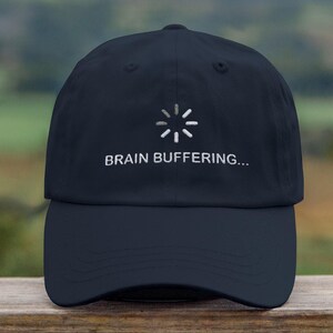 Puede incluir: Una gorra de béisbol azul oscuro con un símbolo de carga blanco y el texto "BRAIN BUFFERING..." bordado en la parte delantera.