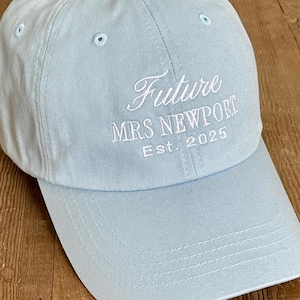 May include: A light blue baseball cap with white embroidery that reads "Future Mrs. Newport Est. 2025".
