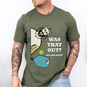 Pu&ograve; includere: Una maglietta verde con un'immagine di un uomo dei cartoni animati che tiene una racchetta da pickleball. Il testo sulla maglietta recita "Was that out? Are you sure?"