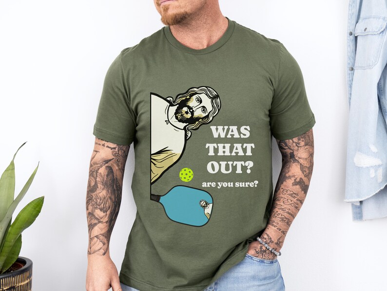 Pu&ograve; includere: Una maglietta verde con un'immagine di un uomo dei cartoni animati che tiene una racchetta da pickleball. Il testo sulla maglietta recita "Was that out? Are you sure?"