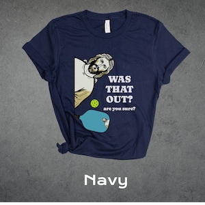 Pu&ograve; includere: Una maglietta blu navy con un'immagine di cartone animato di Ges&ugrave; che gioca a pickleball. Il testo sulla maglietta recita "Was that out? are you sure?"