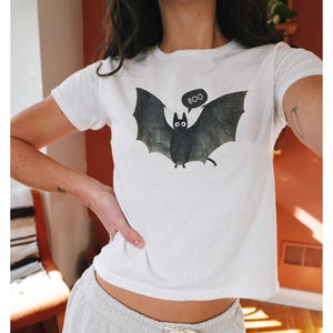 K&ouml;nnte beinhalten: Wei&szlig;es, bauchfreies T-Shirt mit einem schwarzen Fledermaus-Motiv und dem Wort "BOO" in einer Sprechblase.