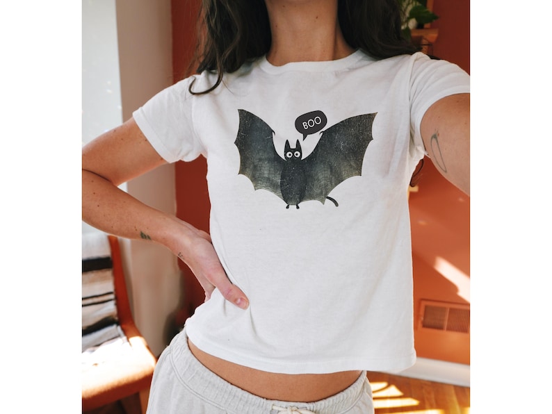 K&ouml;nnte beinhalten: Wei&szlig;es, bauchfreies T-Shirt mit einem schwarzen Fledermaus-Motiv und dem Wort "BOO" in einer Sprechblase.