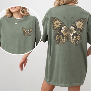 Pu&ograve; includere: Una maglietta oversize verde con un design a farfalla dorata sul retro. La farfalla &egrave; composta da motivi floreali.