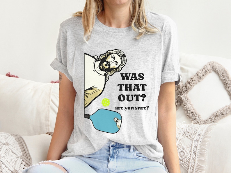 Pu&ograve; includere: Una maglietta grigia con una grafica di Ges&ugrave; che sbircia da dietro una racchetta da pickleball. Il testo sulla maglietta dice "Was that out? Are you sure?"