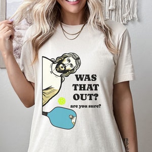 Pu&ograve; includere: Una t-shirt beige con una grafica di Ges&ugrave; che guarda sopra una racchetta da pickleball con il testo "Was that out? are you sure?"