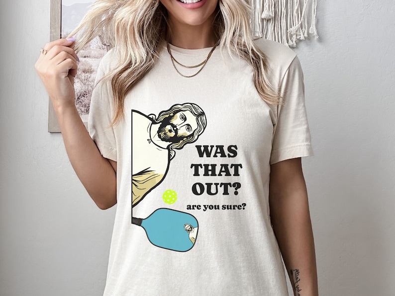 Pu&ograve; includere: Una t-shirt beige con una grafica di Ges&ugrave; che guarda sopra una racchetta da pickleball con il testo "Was that out? are you sure?"