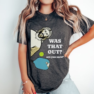 Pu&ograve; includere: Una maglietta grigio scuro con una grafica di Ges&ugrave; che guarda sopra una racchetta da pickleball con il testo "Was that out? Are you sure?"