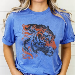 May include: A blue t-shirt featuring a graphic of a tiger's head in black and orange with a distressed effect.