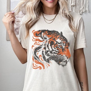 May include: A beige t-shirt featuring a vintage-style orange and black tiger graphic. The tiger's head is facing to the right and has a distressed look.