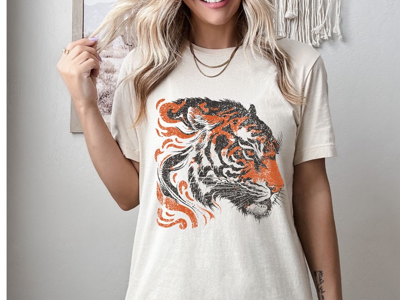 May include: A beige t-shirt featuring a vintage-style orange and black tiger graphic. The tiger's head is facing to the right and has a distressed look.