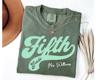 May include: A green t-shirt with a mint green graphic that says "Fifth Grade" in a retro font. The teacher's name, "Mrs. Williams", is written below the graphic.