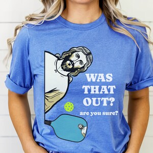 Pu&ograve; includere: Una maglietta blu con una grafica di Ges&ugrave; che sbircia da dietro una racchetta da pickleball. Il testo sulla maglietta recita "Was that out? Are you sure?"