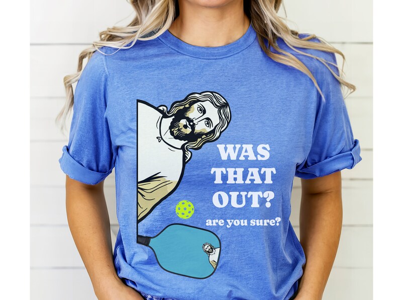 Pu&ograve; includere: Una maglietta blu con una grafica di Ges&ugrave; che sbircia da dietro una racchetta da pickleball. Il testo sulla maglietta recita "Was that out? Are you sure?"