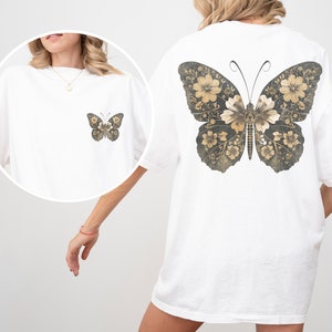 Pu&ograve; includere: Maglietta bianca oversize con un grande disegno di farfalla floreale sul retro. La farfalla &egrave; in tonalit&agrave; di marrone e oro.