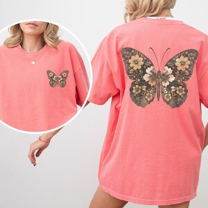 Pu&ograve; includere: Una maglietta oversize color corallo rosa con un grande disegno di farfalla floreale sul retro. La farfalla &egrave; marrone e dorata con dettagli intricati.