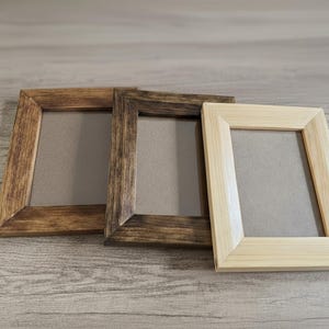 May include: Three wooden picture frames in varying shades of brown and natural wood. The frames are rectangular with a simple, clean design. The frames are arranged side by side on a light wood-grain surface.