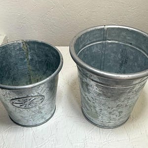 May include: Two galvanized metal planters, one larger than the other, with a distressed finish. The smaller planter has a decorative design on the side.