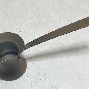 May include: A small, antique-style metal coffee pot with a long, flat handle. The pot is a dark, muted bronze color, and the handle has a hole at the end. This type of pot is often used for making Turkish coffee.