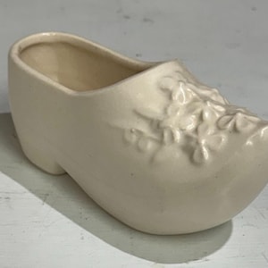 May include: A cream-colored ceramic clog, a traditional Dutch shoe, with a floral design on the toe. The shoe is empty inside and sits on a white surface. The clog is a decorative item.