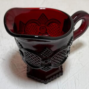 May include: A ruby red glass creamer with a decorative floral and geometric pattern. The creamer has a handle and a spout for pouring. The base is square and the creamer is approximately 10 cm tall.