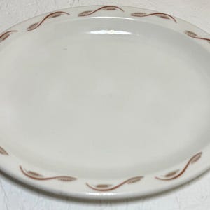 May include: An oval, off-white ceramic serving platter with a brown leaf and vine design around the rim. The plate is approximately 12 inches long and 9 inches wide, with a smooth, glossy surface.