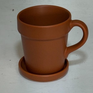 May include: A terracotta-colored mug with a matching saucer. The mug has a rounded handle and a design that mimics a flower pot. The mug and saucer are made of ceramic and are a solid, earthy tone.