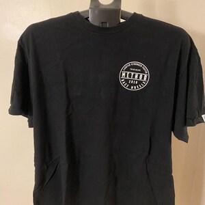 May include: Black t-shirt with a crew neck and short sleeves. A white circular logo on the upper left chest reads "METHOD 2010 RACE WHEELS" with additional text around the perimeter. The shirt is hanging on a black hanger.