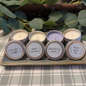 May include: A set of eight small, round, silver-colored candles in tins, arranged on a rectangular ceramic tray. Each candle has a white label with a different scent name: Pumpkin Muffin, Apple Harvest, Elderberry Nectar, and Maple Apple Pie.
